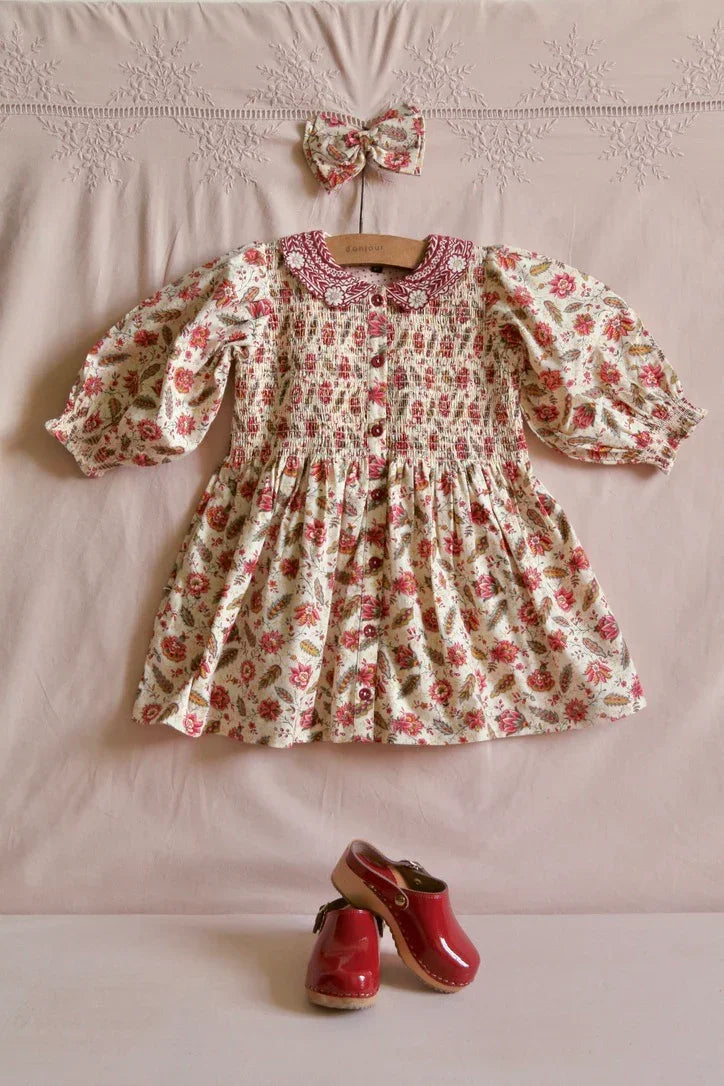 Floral smocked baby dress with peter pan collar, matching hair bow, and red clogs on pink linen