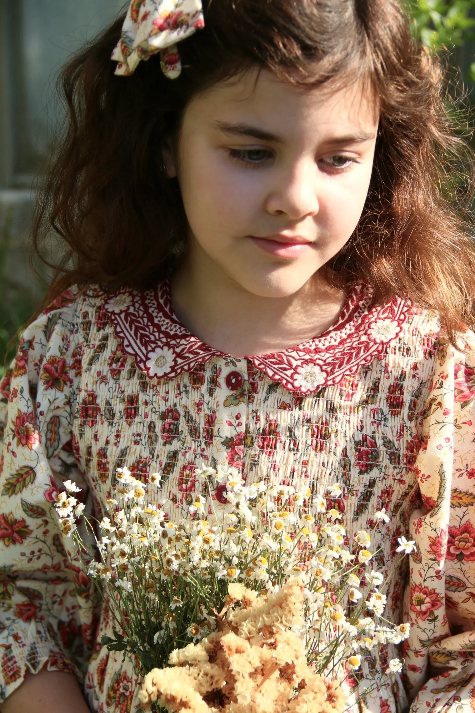 Girl in vintage floral print dress and hair bow holding chamomile bouquet outdoors