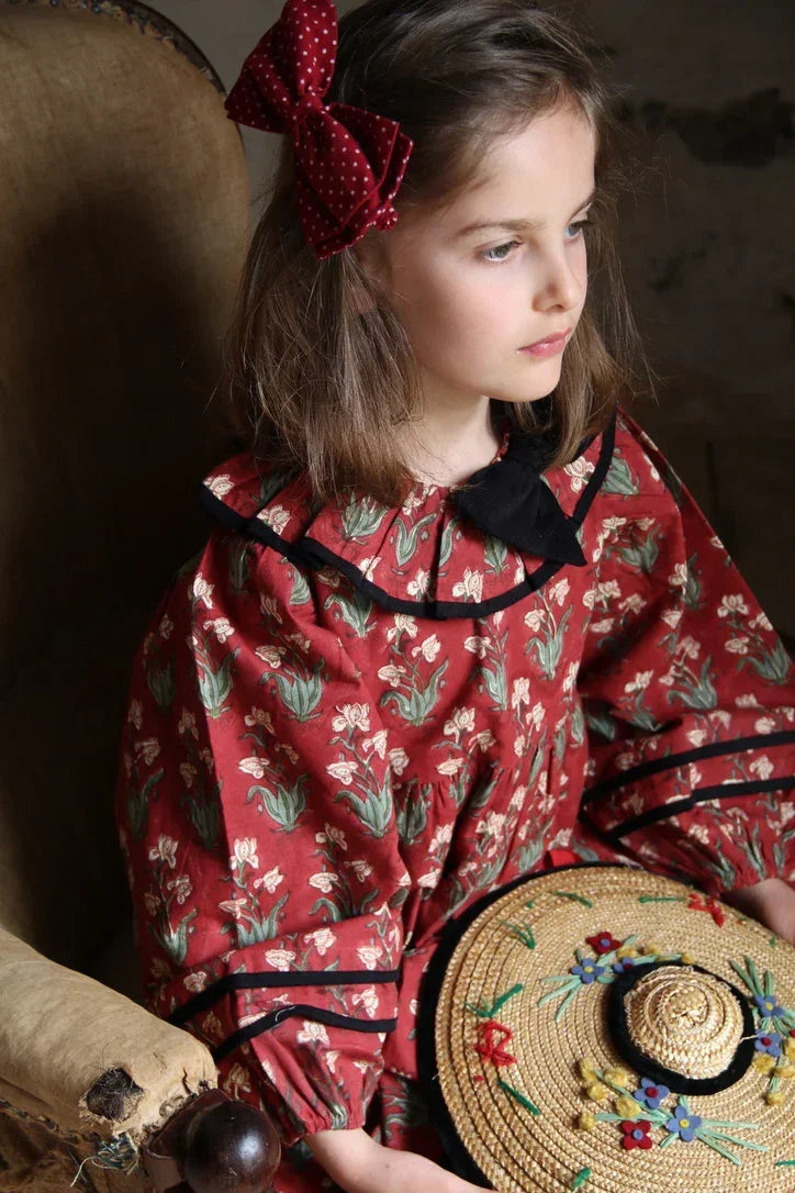 Girl in a red floral print dress with black trim, red polka dot hair bow, holding an embroidered straw hat.