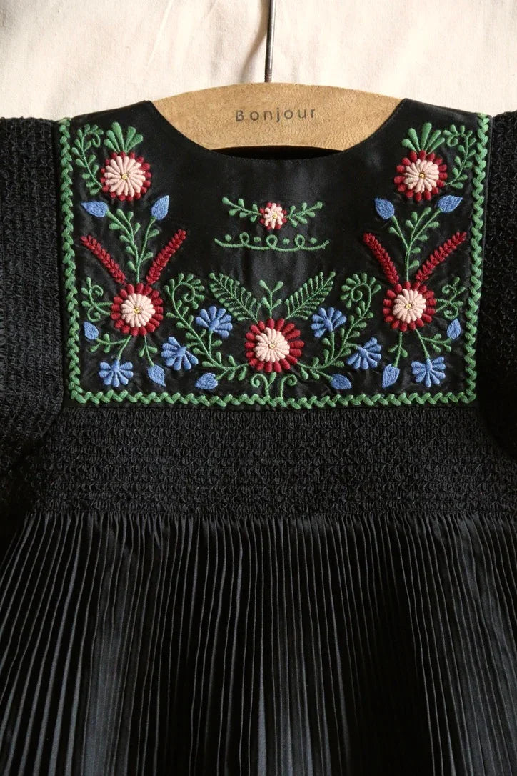 Black pleated children's dress with colorful floral embroidery on the yoke, hanging on a hanger.