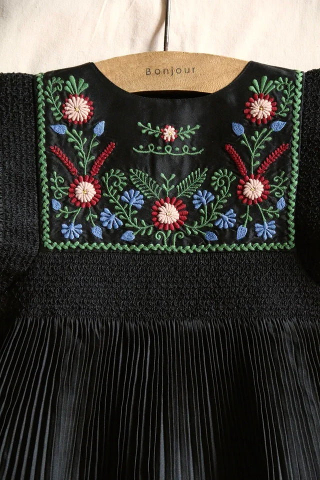 Black pleated children's dress with colorful floral embroidery on the yoke, hanging on a hanger.