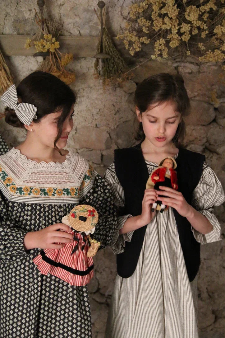 Two girls in vintage-style childrenswear holding dolls, standing by a rustic stone wall with dried flowers.