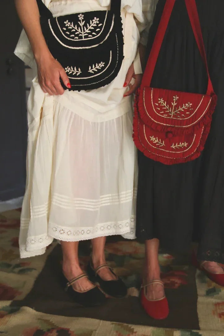Two women in vintage style skirts holding embroidered crossbody bags, one black and one red