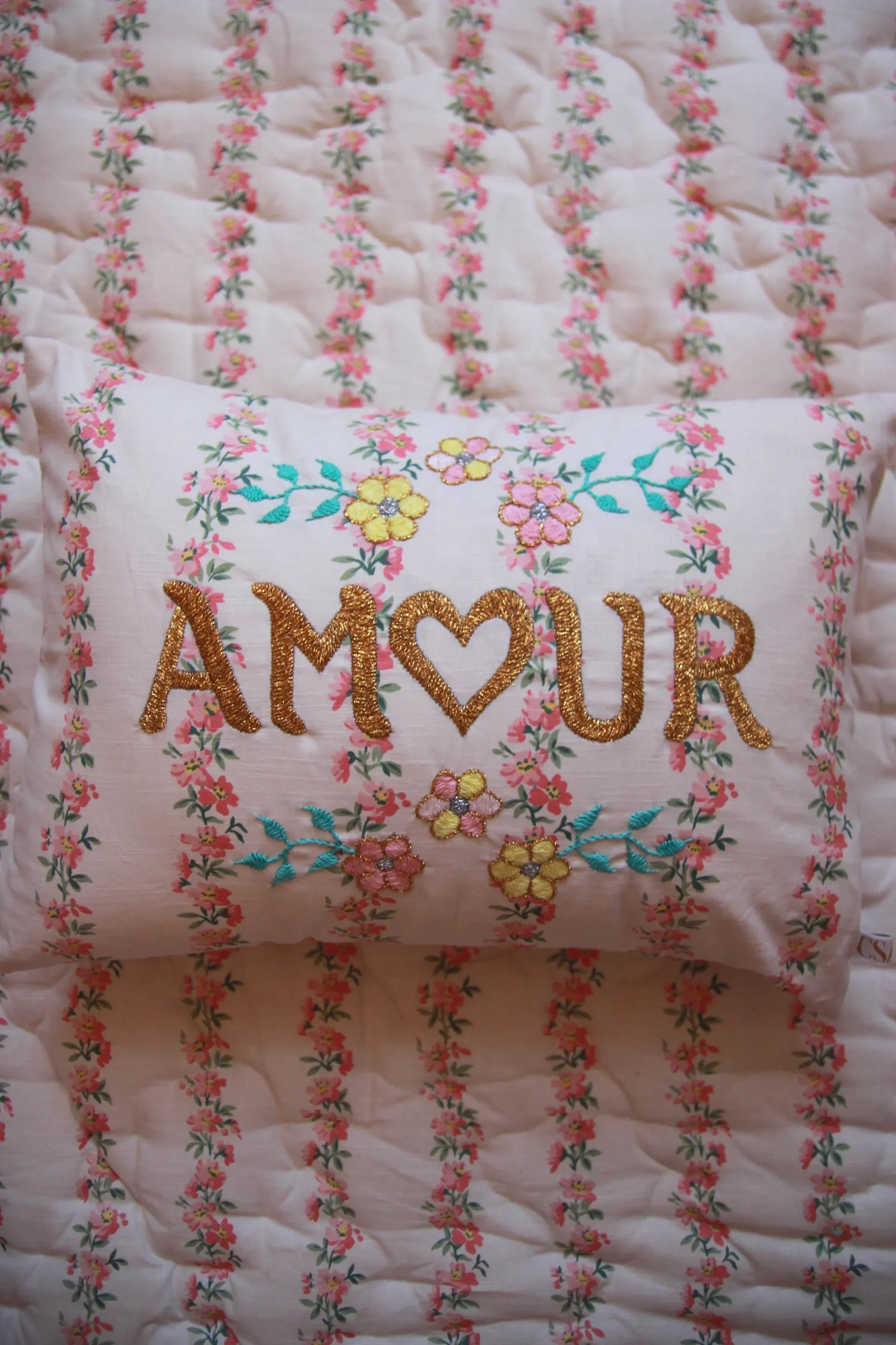 Floral embroidered pillow with 'AMOUR' text on pink flower-printed bedding.
