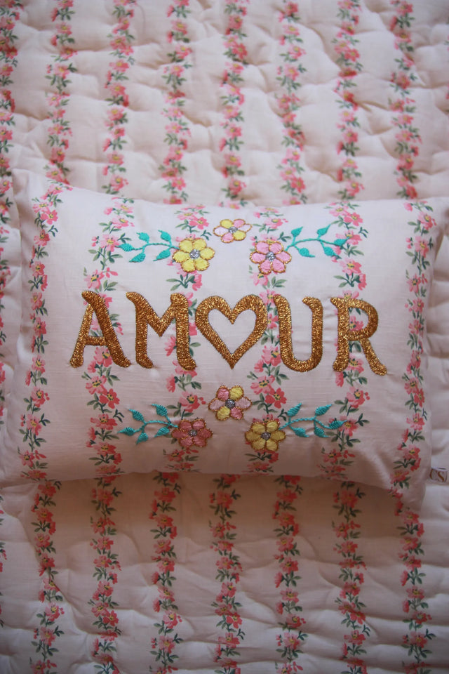 Floral embroidered pillow with 'AMOUR' text on pink flower-printed bedding.