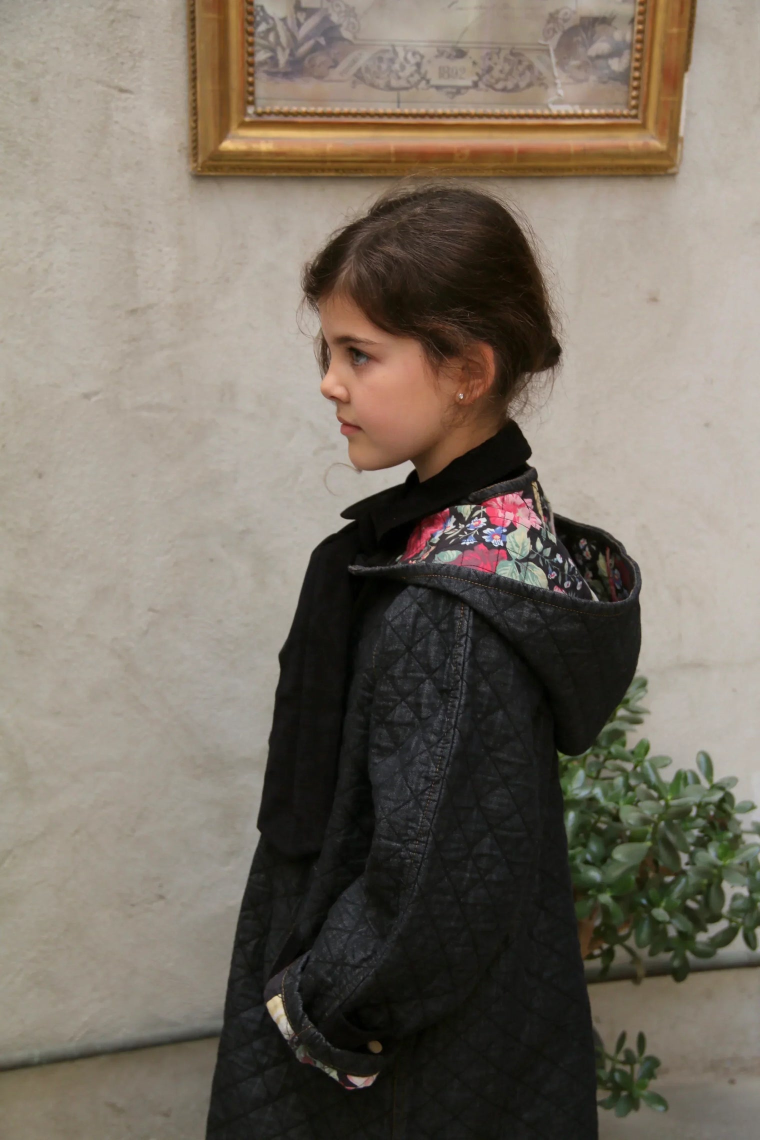 Girl wearing quilted black children's coat with floral patterned hood, side profile indoors