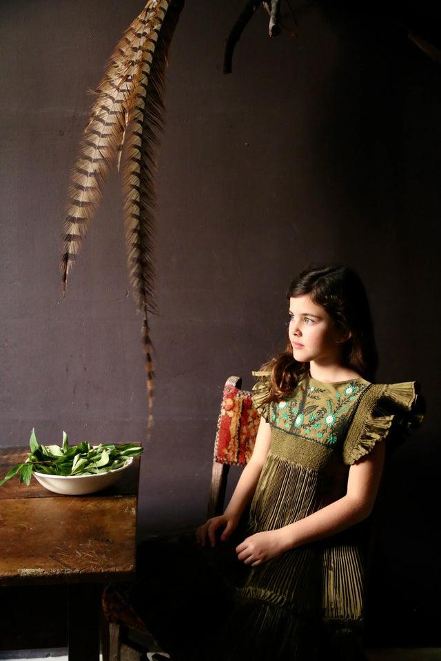Girl in olive green embroidered dress sitting by a table with green leaves, dark background