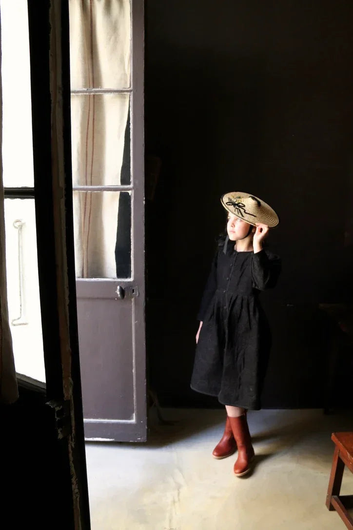 Child in a black dress and straw hat standing indoors by a door, wearing brown boots