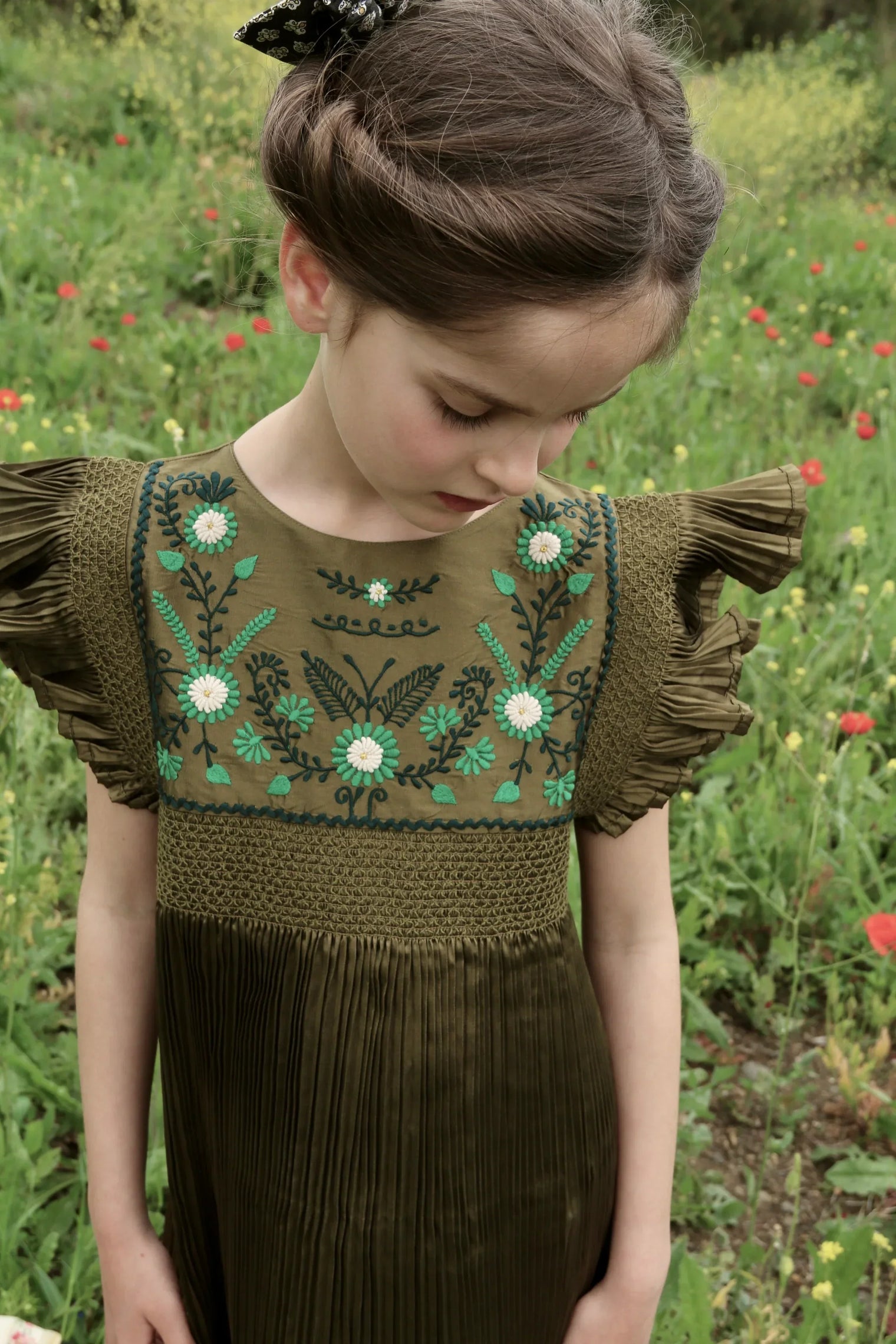Girl in olive green embroidered dress with pleated sleeves, standing in a wildflower field