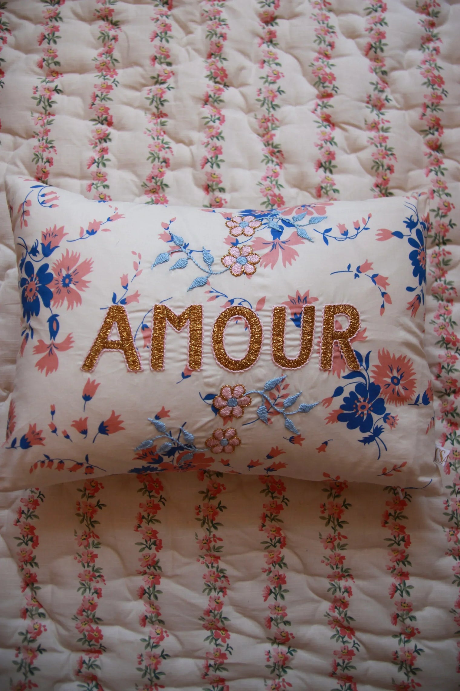 Decorative floral embroidered cushion with 'AMOUR' text on patterned quilt bedding
