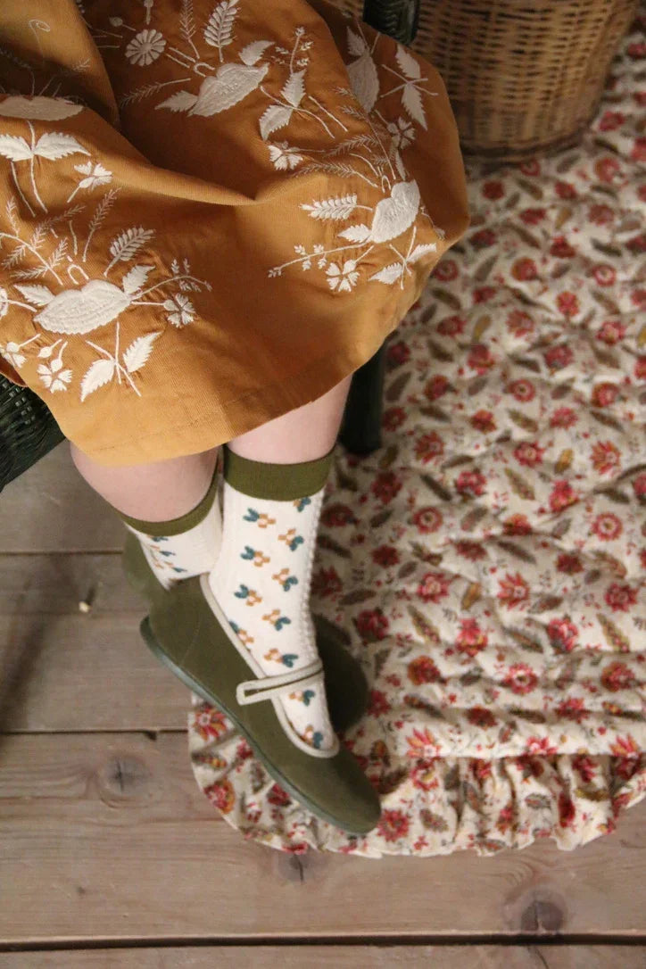 Child in yellow embroidered dress, floral socks, and green Mary Jane shoes on patterned rug