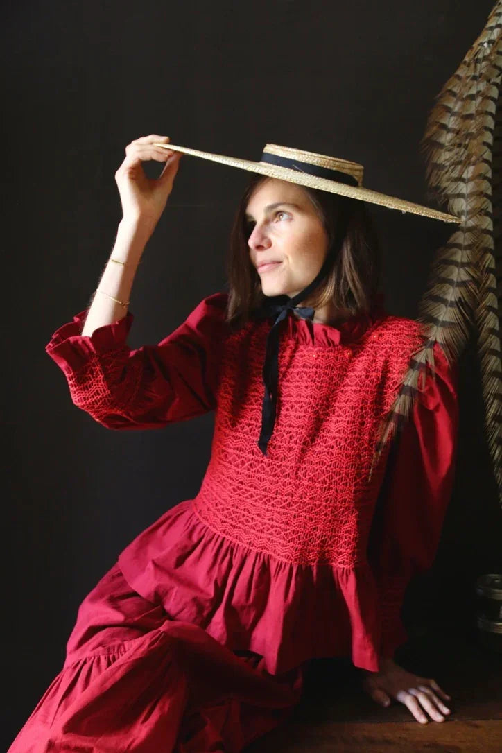Woman in red smocked dress with black ribbon, wearing straw hat, feathers in background