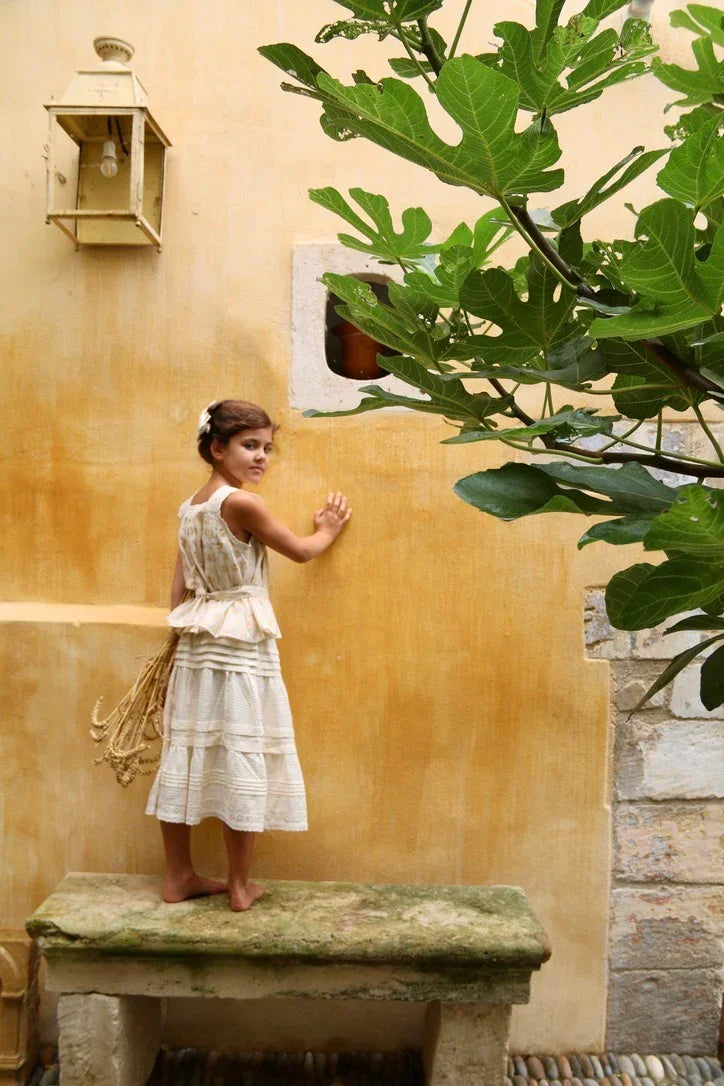 Girl in vintage cream dress standing barefoot on stone bench by yellow wall with fig tree.