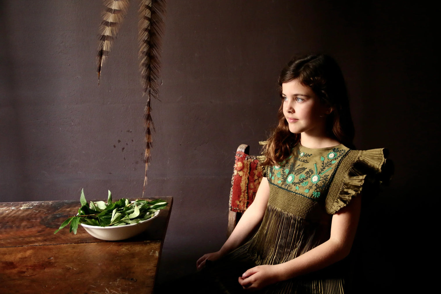 Girl in an embroidered green dress with ruffle sleeves sitting beside a rustic table with a bowl of greens, vintage setting.