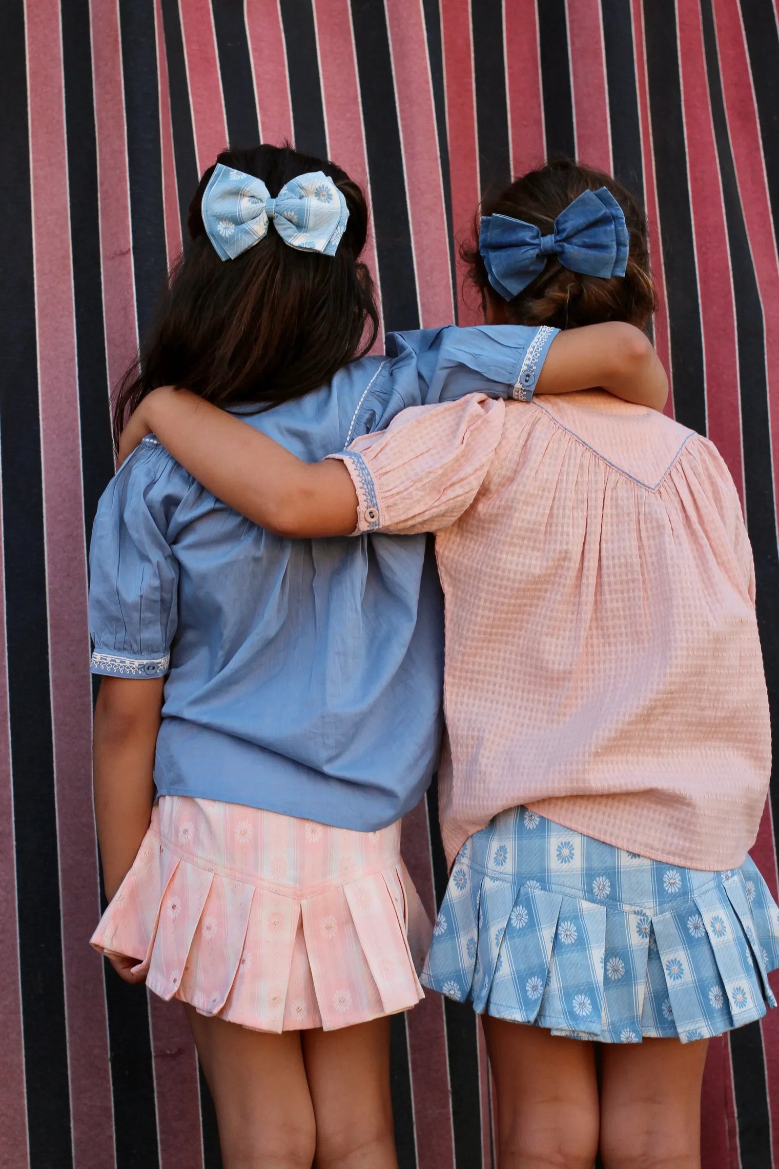 Two girls in pastel childrenswear with pleated skirts and bows, arms around each other