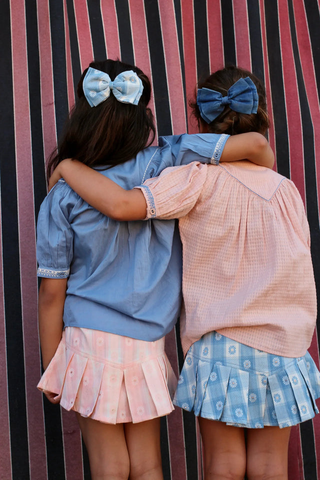 Two girls in pastel childrenswear with pleated skirts and bows, arms around each other