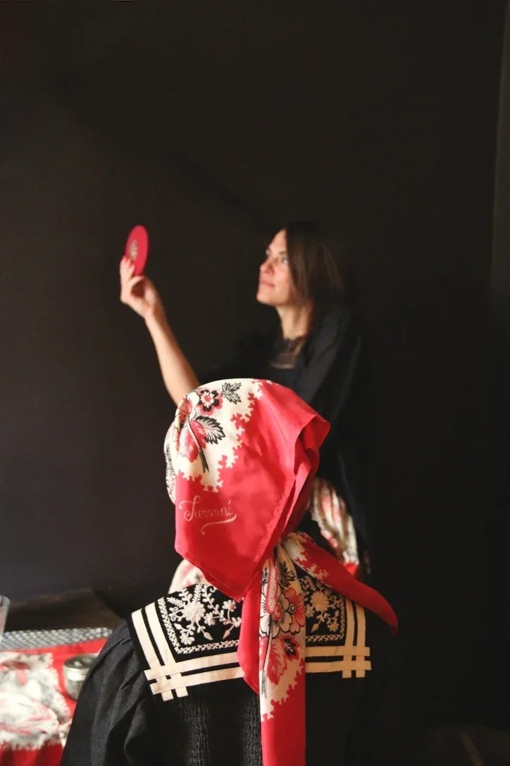 Woman in black dress and floral red headscarf sitting indoors with another woman holding a red mirror