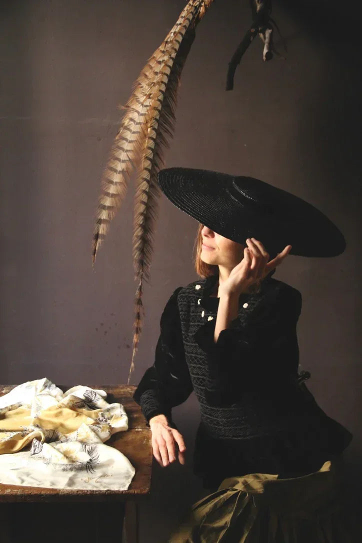Woman in black wide-brim hat and vintage blouse sitting by table with patterned linens