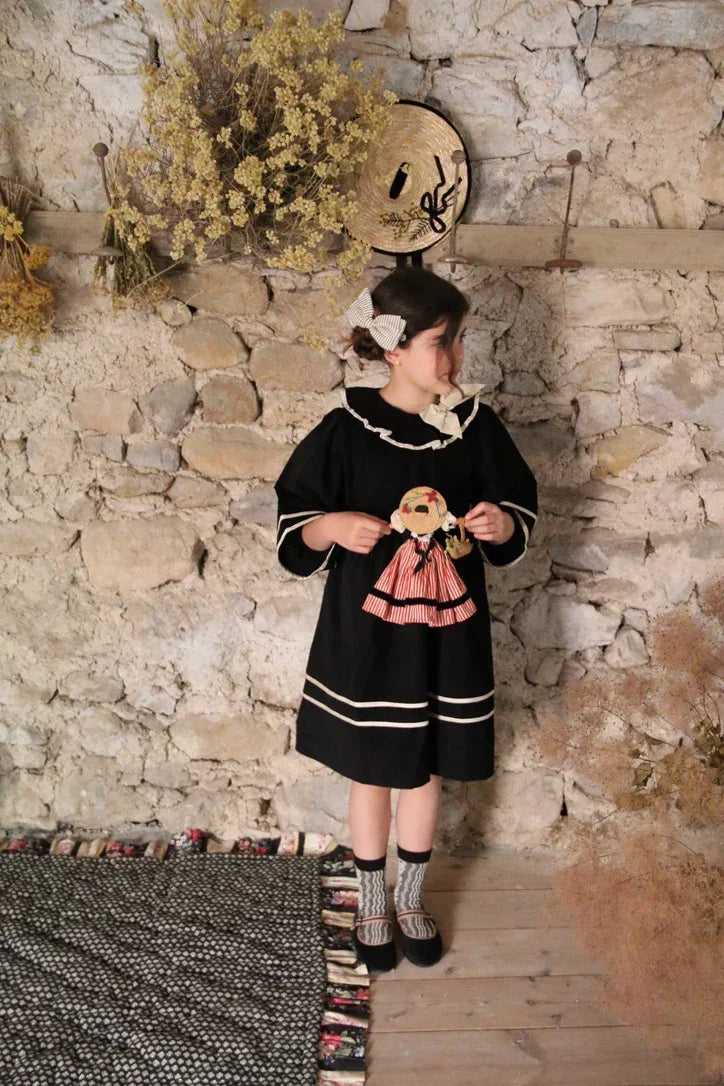 Girl in black vintage-inspired dress with doll, standing by rustic stone wall and dried flowers