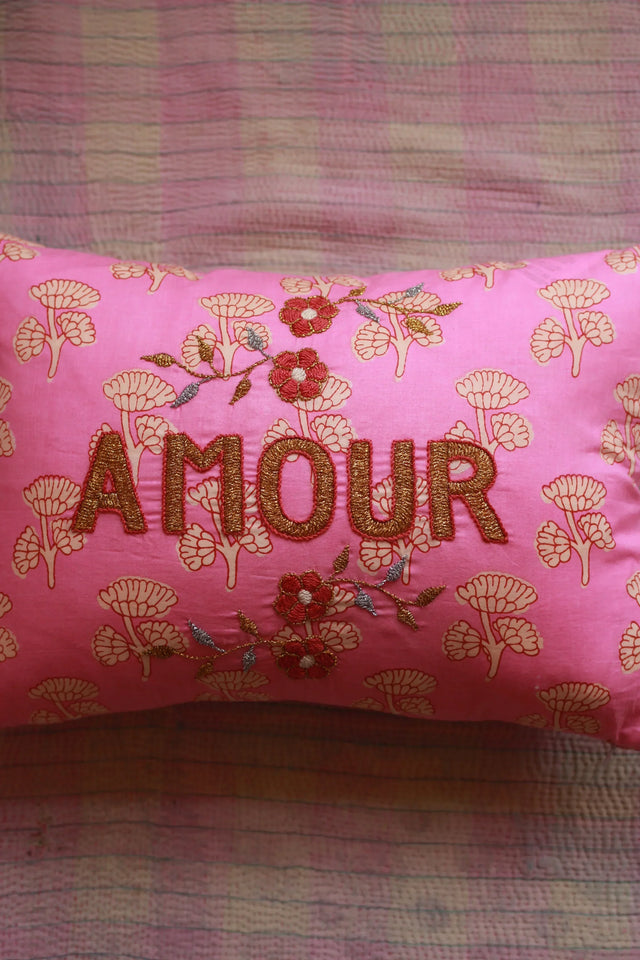 Pink decorative pillow with floral pattern and embroidered 'AMOUR' text on patterned fabric