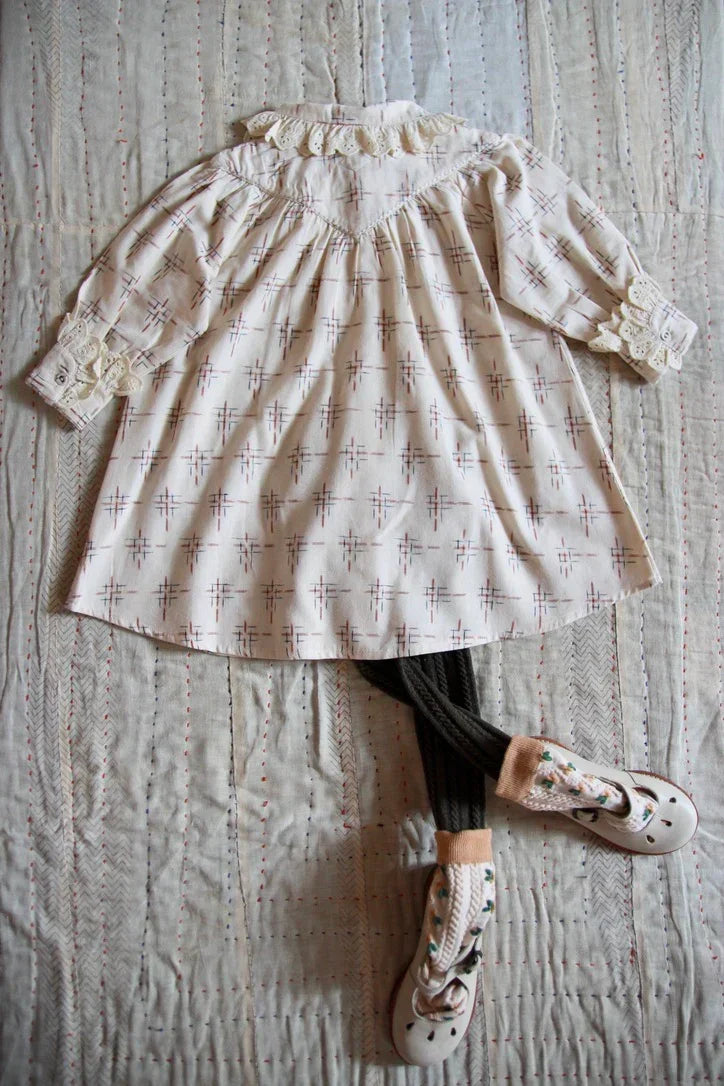 Vintage-inspired children's dress with lace collar, patterned tights, and embroidered shoes on quilted fabric.