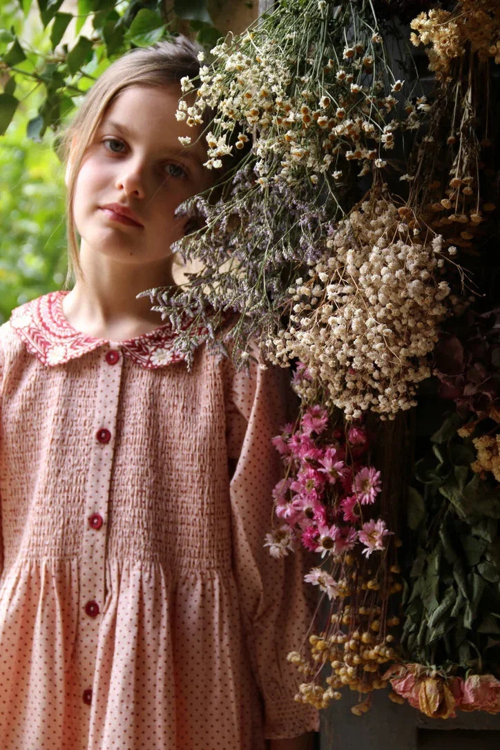 Girl in vintage pink polka dot dress with red buttons beside hanging dried flowers