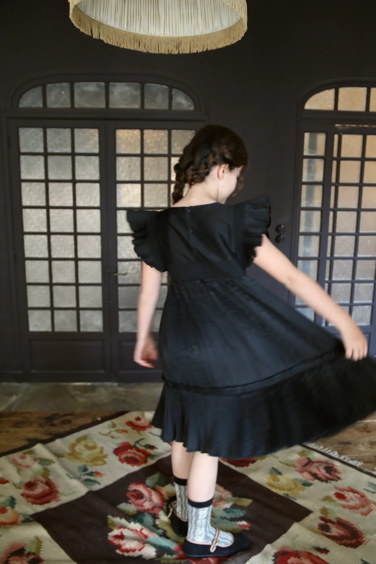 Girl in black ruffled dress twirling on floral rug in vintage room with glass doors