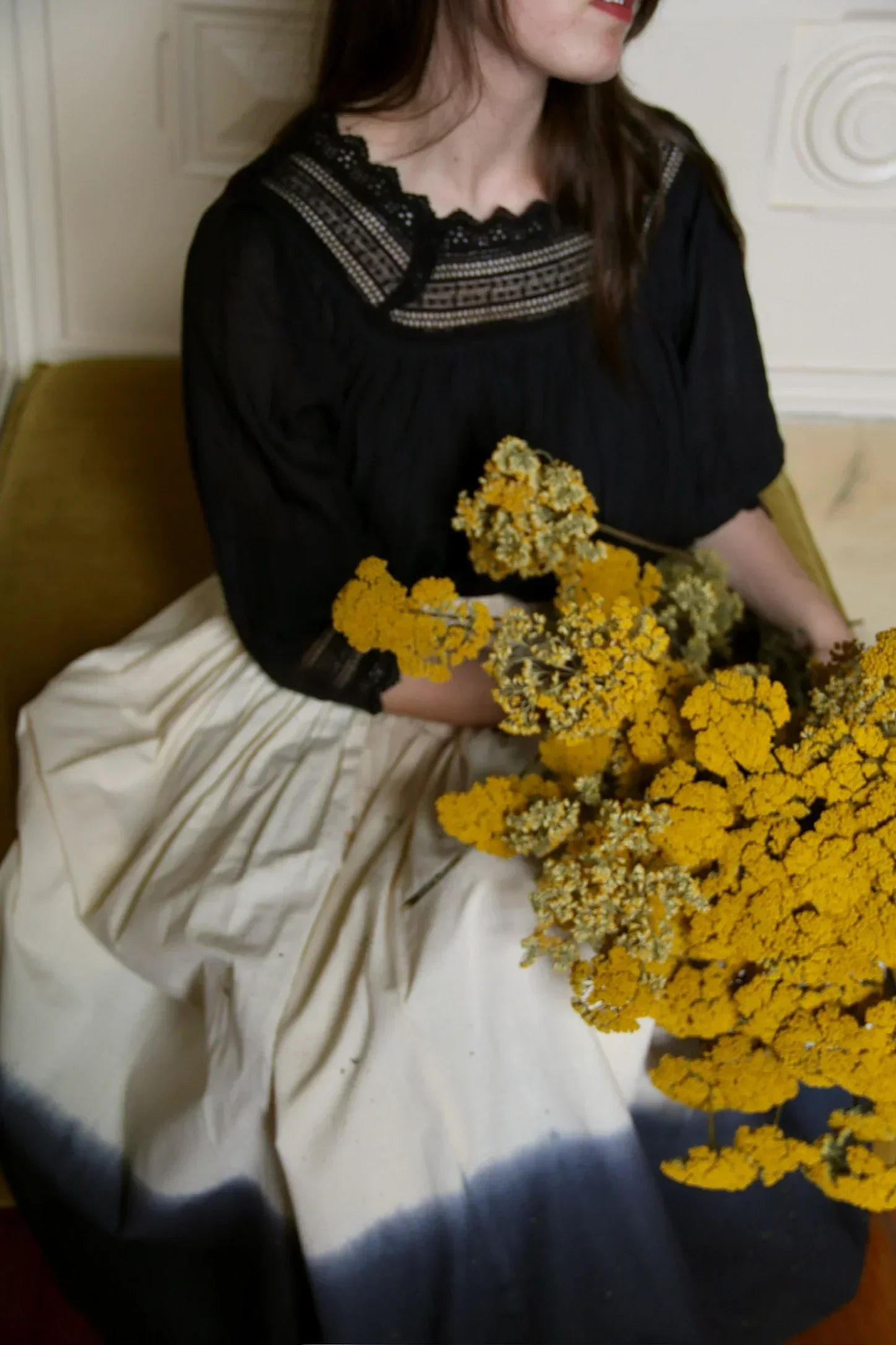 Woman in black lace blouse and white skirt holding yellow flowers indoors