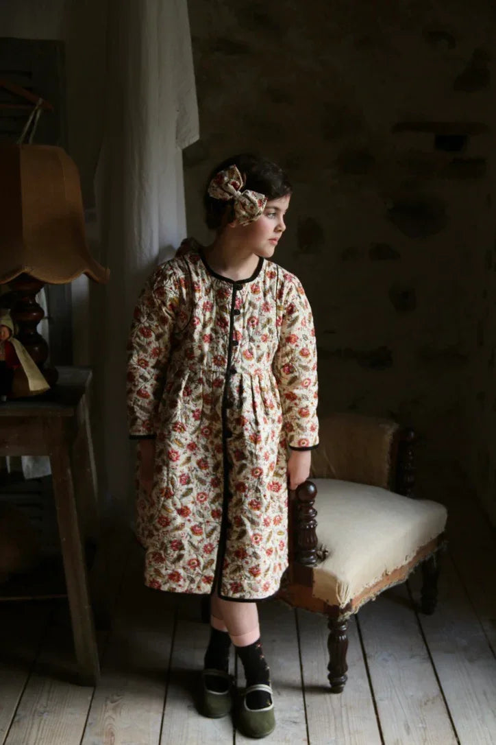 Child wearing floral print quilted dress and matching hair bow, standing in rustic room
