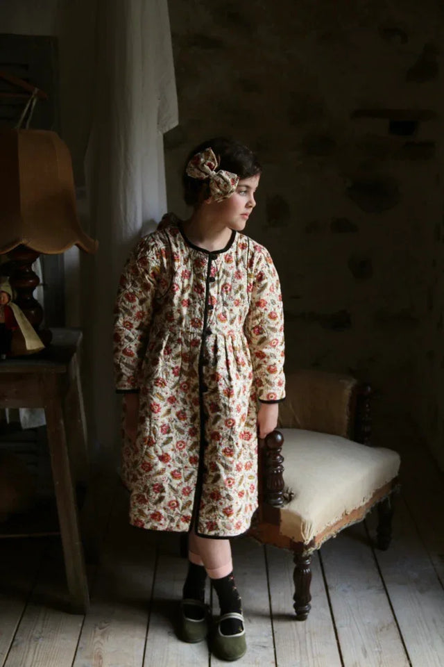 Child wearing floral print quilted dress and matching hair bow, standing in rustic room