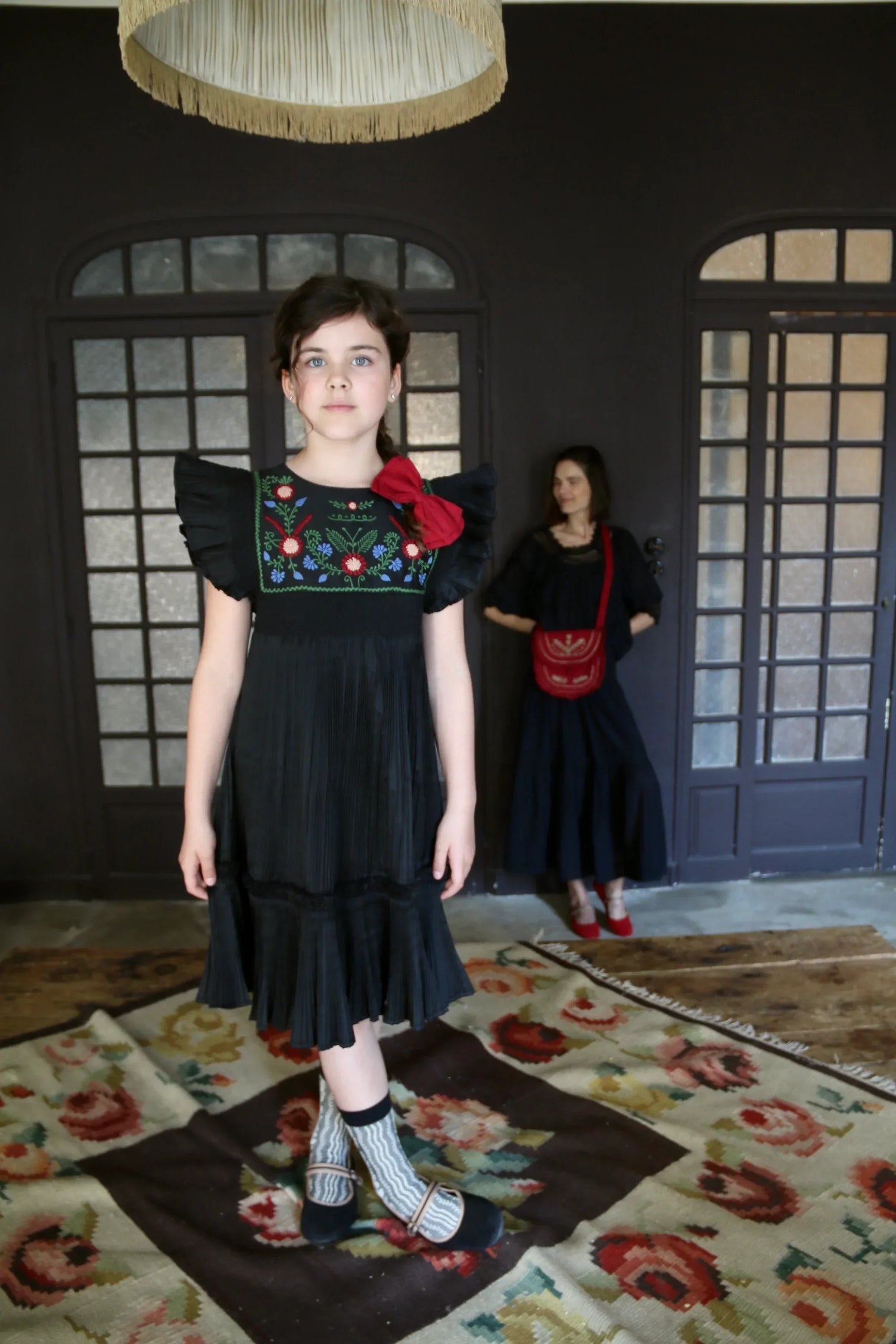 Girl in black ruffled dress with floral embroidery and red bow, standing on floral rug indoors