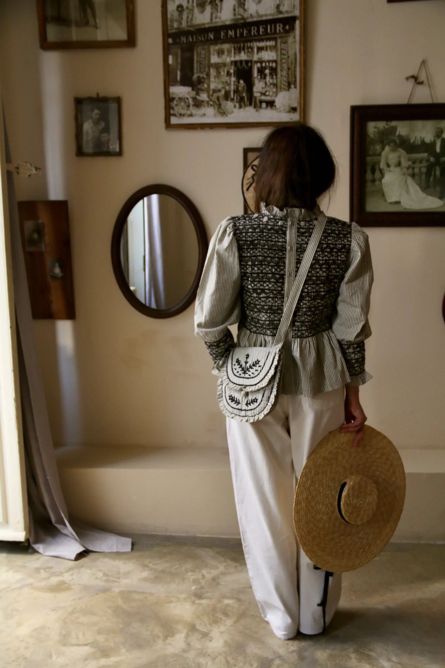 Woman in embroidered top and white pants holding straw hat, vintage wall decor background