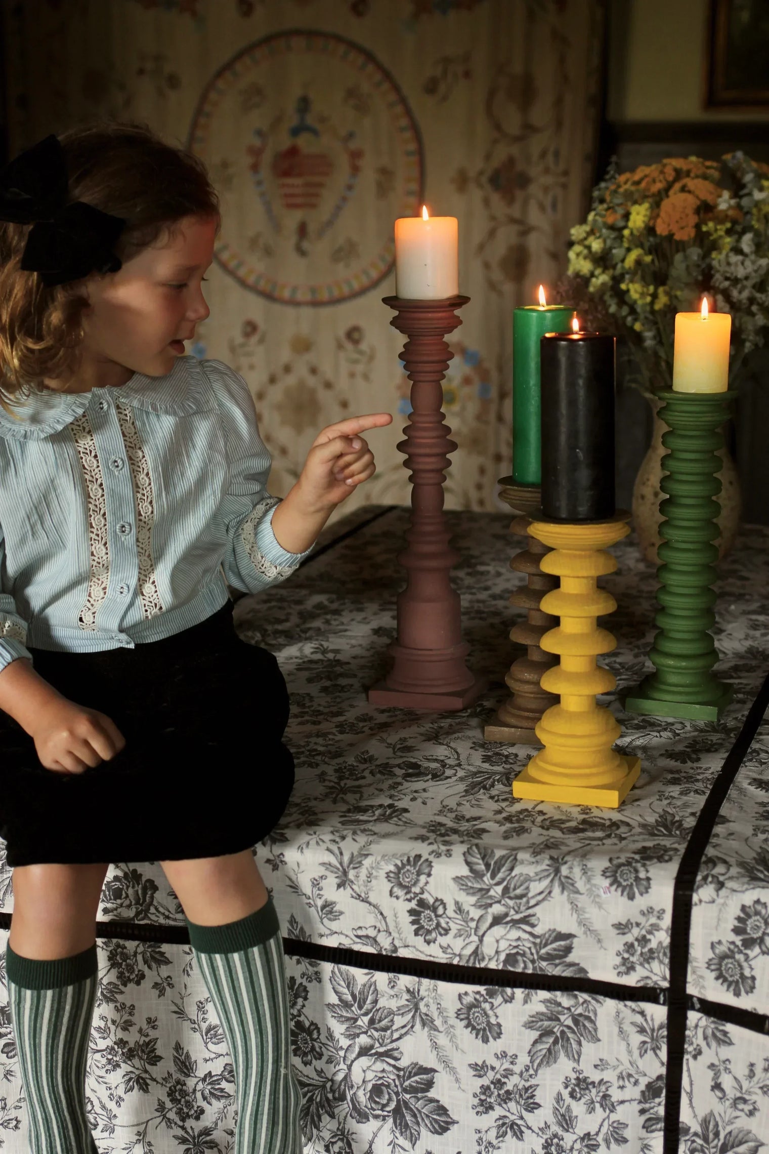 Girl in vintage blouse points at colorful pillar candles on patterned tablecloth