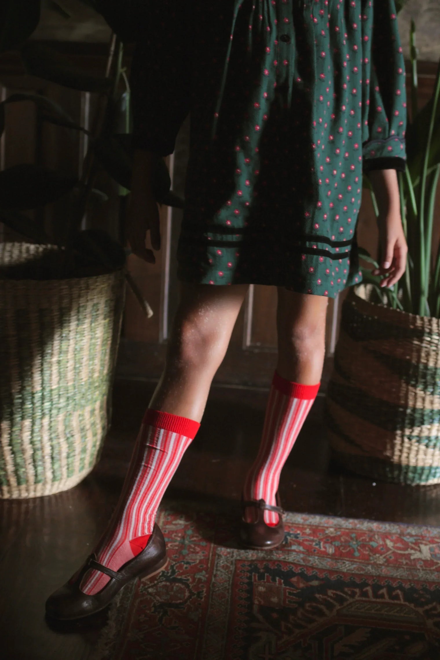 Child in green dress with red pattern, striped knee socks, and brown shoes on patterned rug