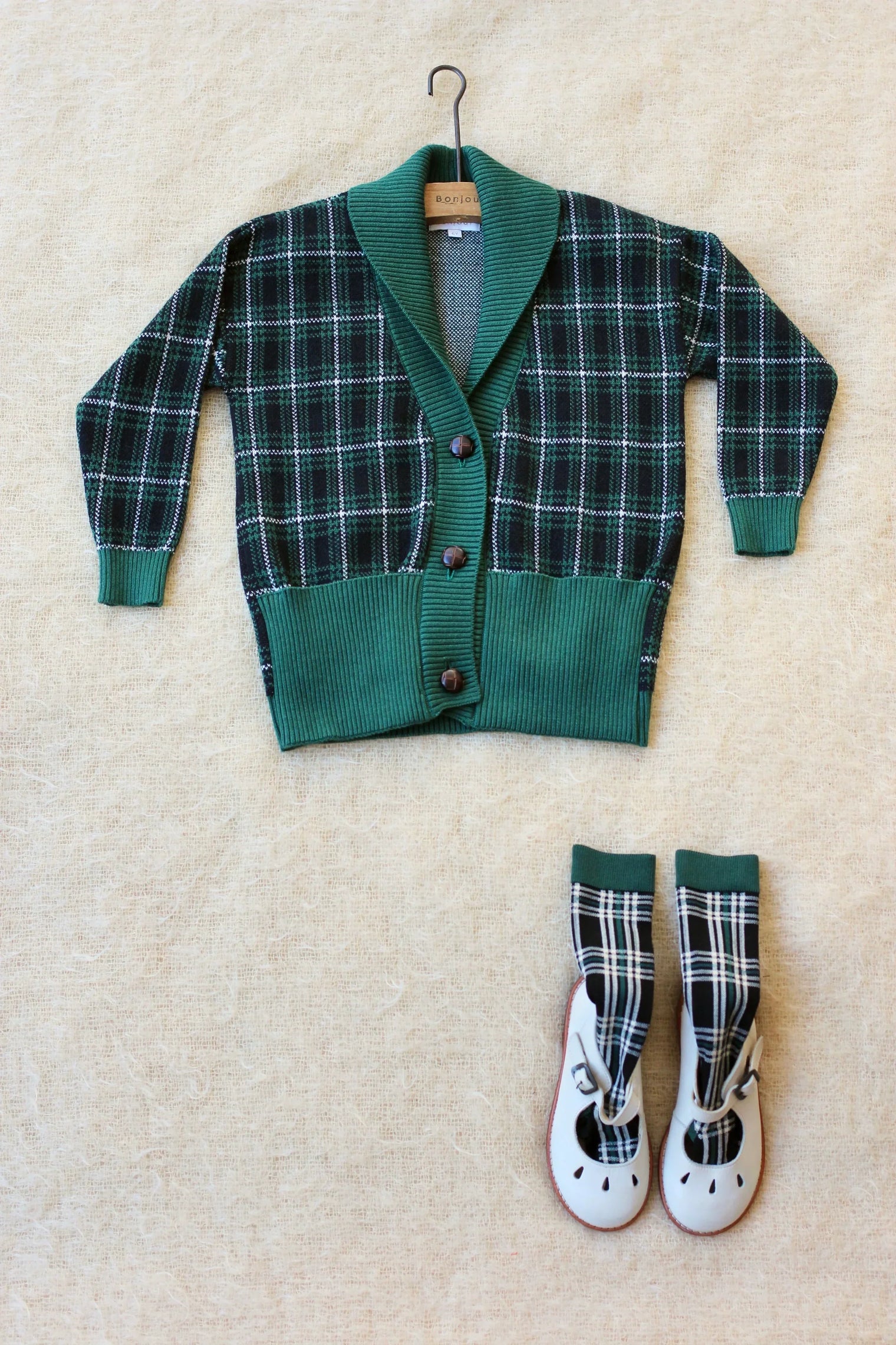 Green plaid children's cardigan with matching socks and white shoes on beige fabric