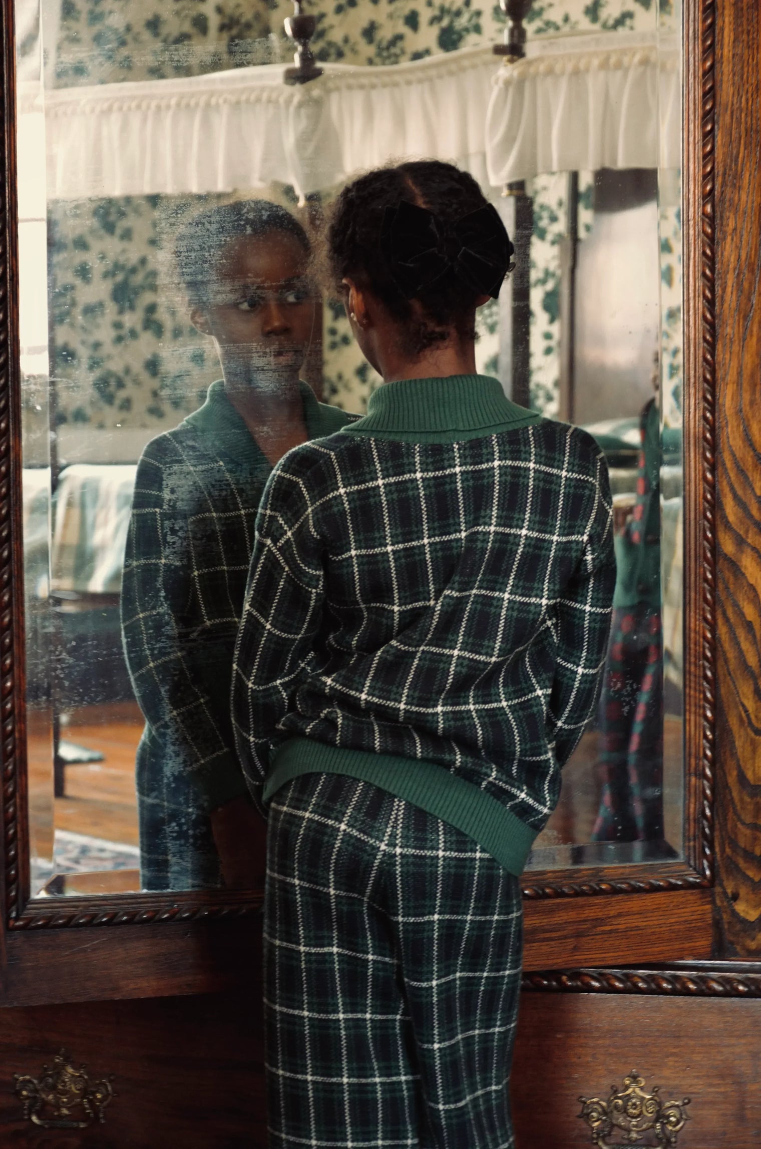 Child in green checkered pajamas with hair bow looking in antique mirror in vintage bedroom