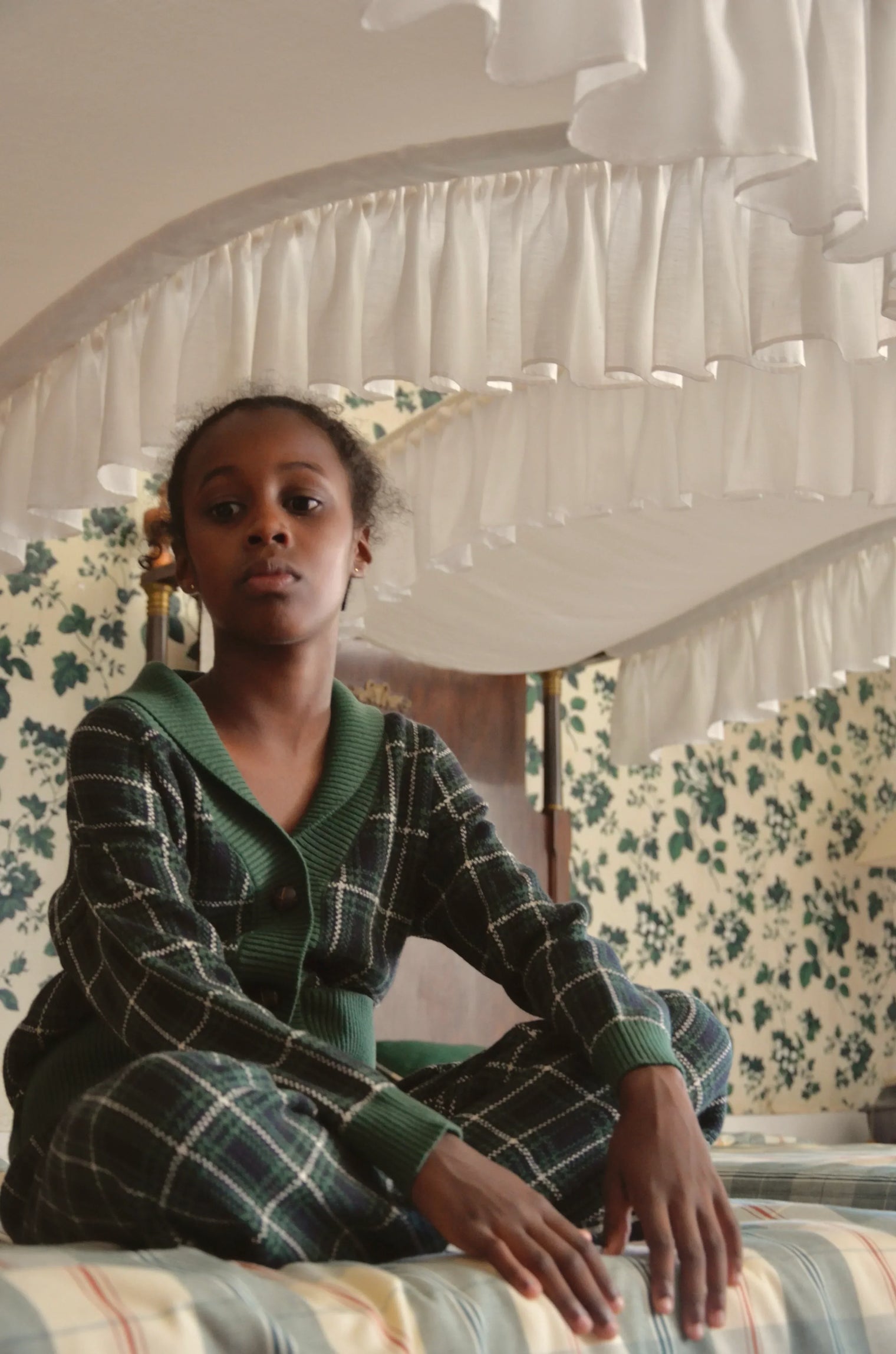 Child in green plaid loungewear sitting on bed with ruffled canopy, floral wallpaper background