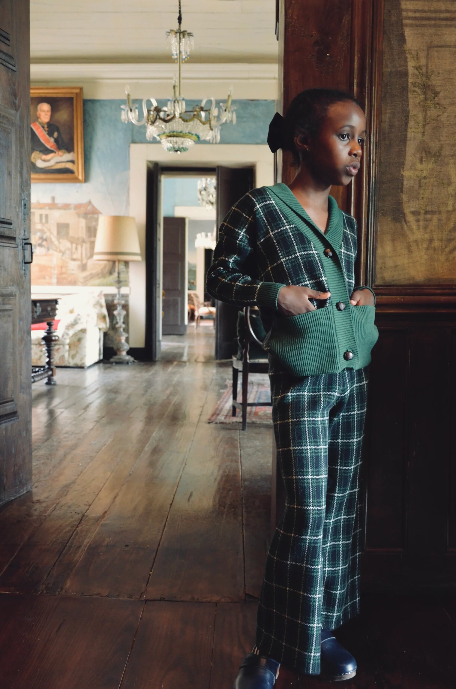 Child in green plaid knit cardigan and pants standing in a vintage, elegant room