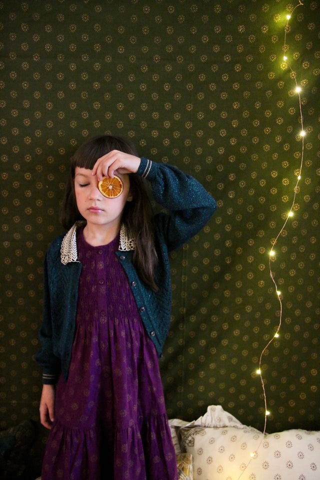 Girl in purple dress and blue cardigan holds dried orange slice, standing by patterned green wall with string lights.