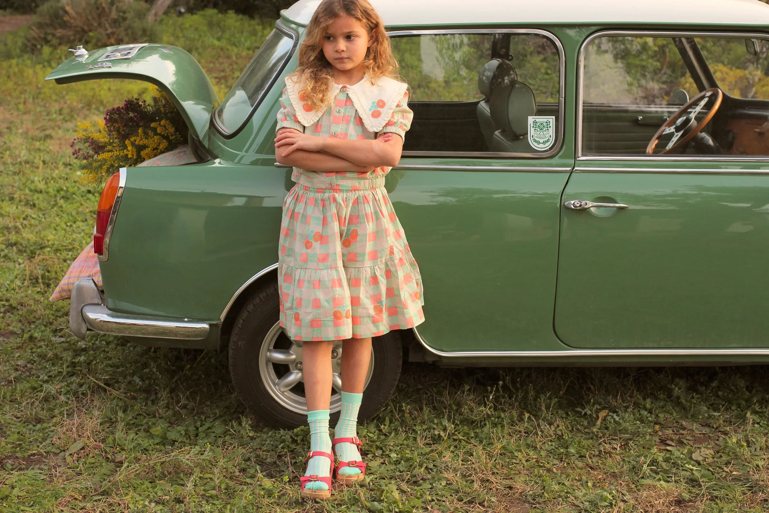 Girl in vintage-style pastel check dress with floral accents standing by green classic car outdoors