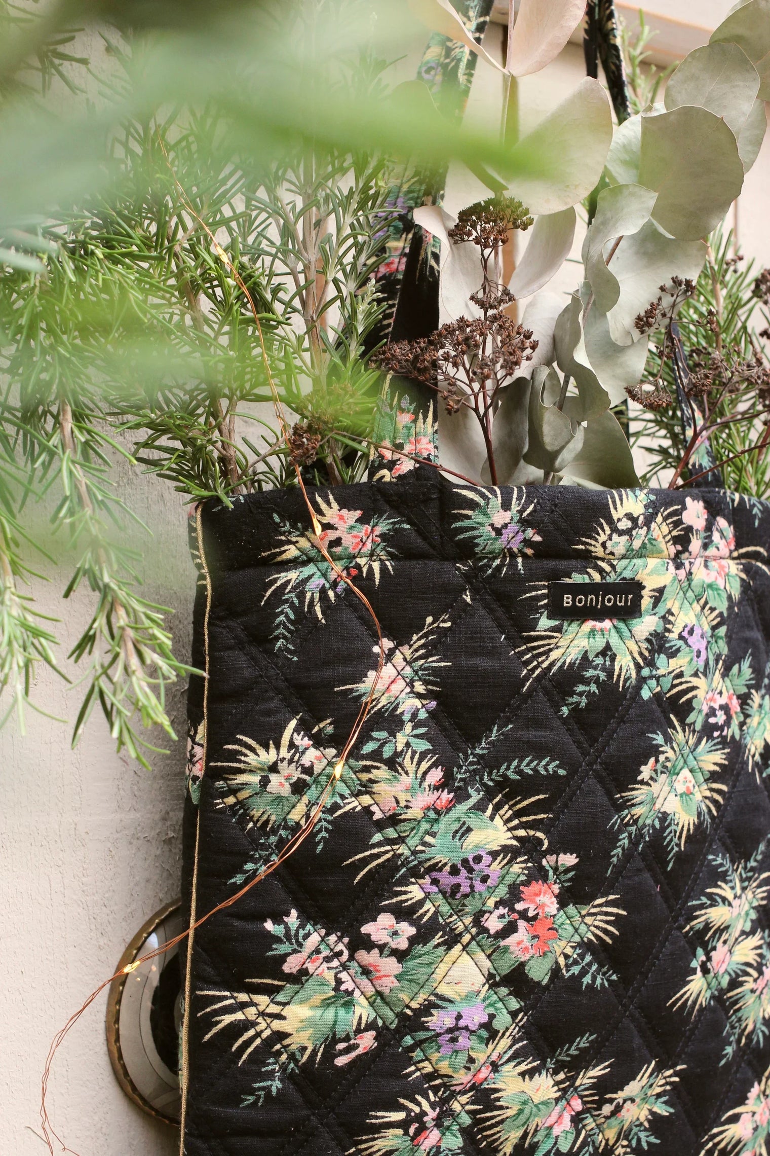 Black quilted floral tote bag with greenery and dried flowers, hanging on a wall