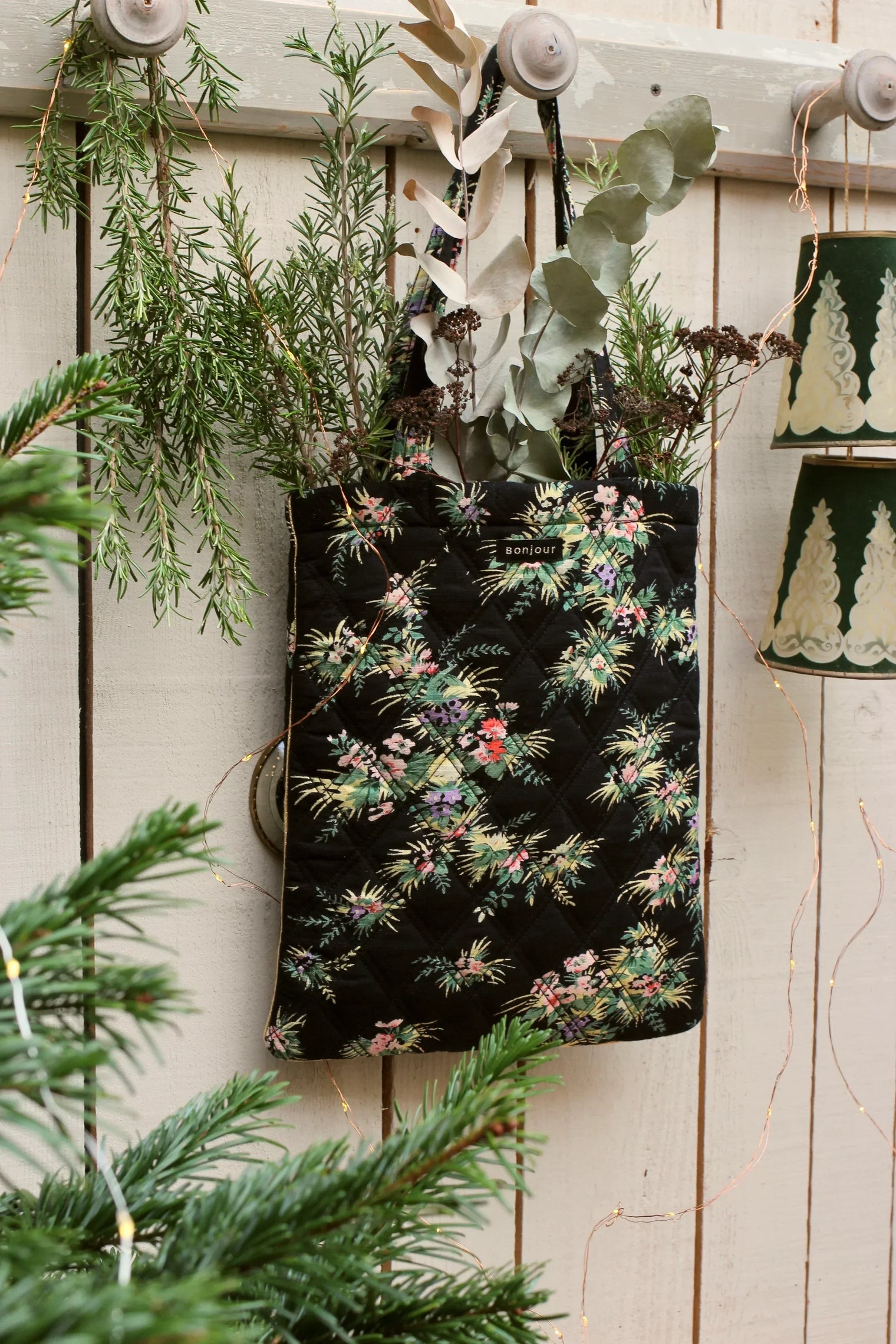 Quilted floral tote bag with botanical print hanging on a white wooden wall, filled with greenery