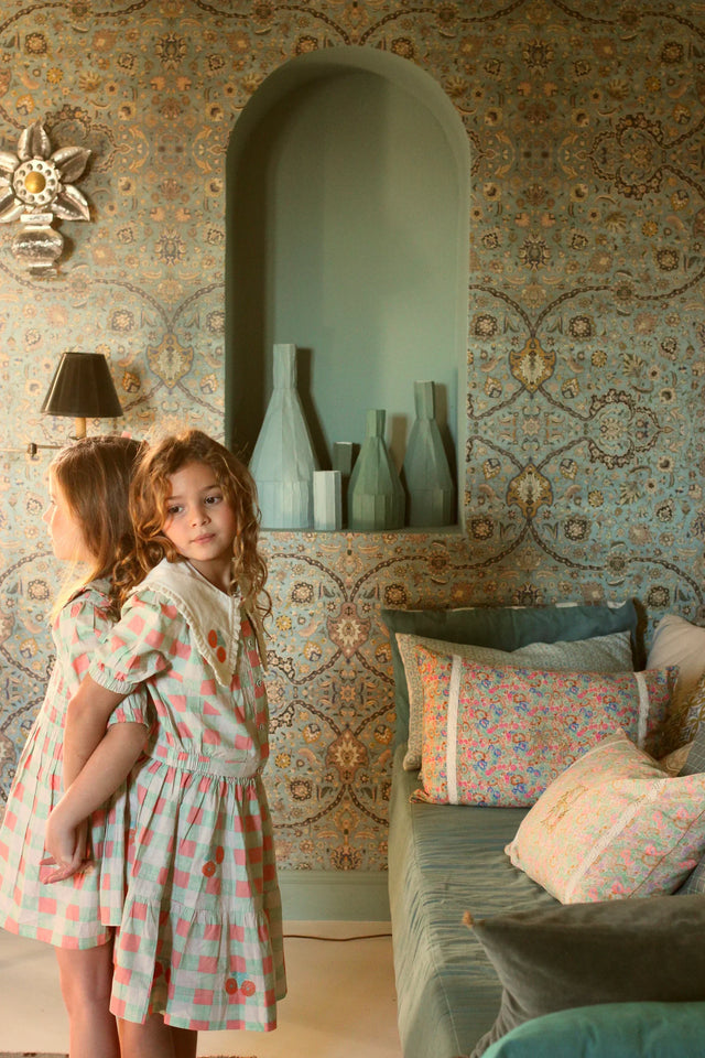 Two young girls in matching checkered dresses stand in a vintage-style living room with patterned wallpaper, decorative throw pillows, and vases on a wall shelf.
