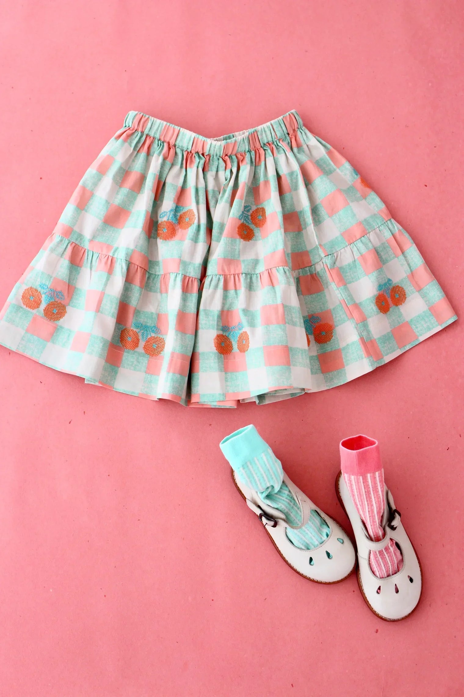Girls’ pastel gingham skirt with floral embroidery, kids’ shoes and socks on pink background