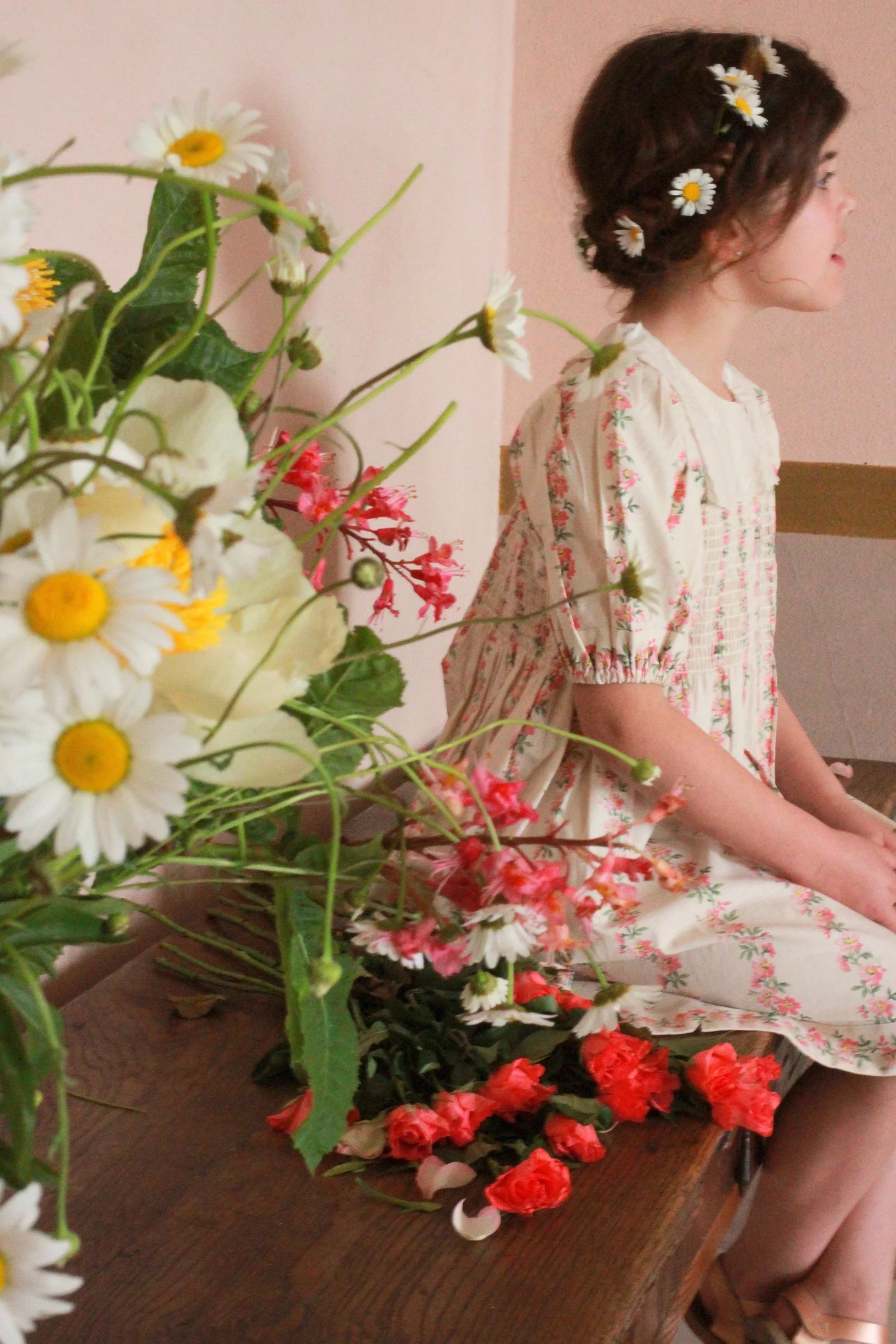 Girl in vintage floral dress with flower crown sitting by bouquet of fresh daisies and roses