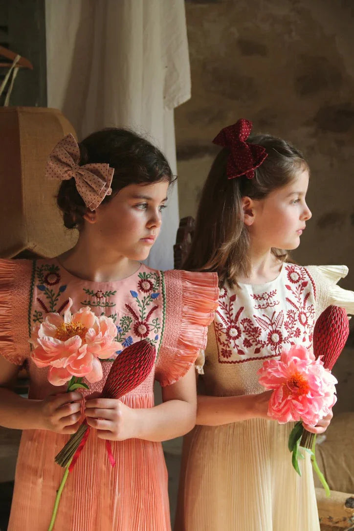 Two girls in embroidered dresses with floral prints, holding flowers, wearing hair bows.