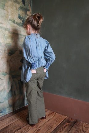Girl in blue gingham blouse and olive embroidered pants standing on wooden floor indoors