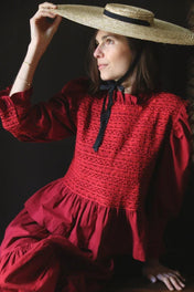 Woman in a red smocked dress with puff sleeves and straw hat with black ribbon