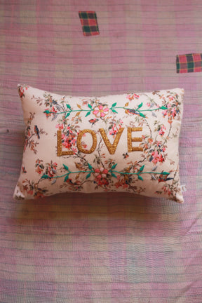 Embroidered floral cushion with gold 'LOVE' text on a pastel quilted bedspread