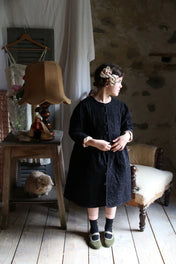Girl in a black quilted dress with floral hair bow standing in rustic vintage room with lamp and cat
